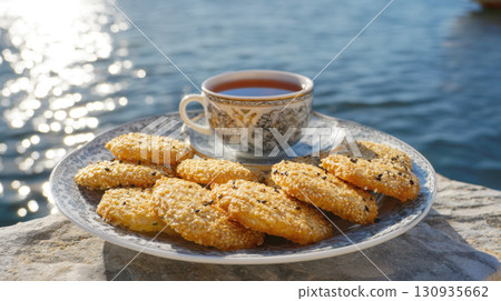 A plate of delicious golden sesame seed cookies with a cup of hot tea on a stone table, set against a backdrop of a sun-drenched, sparkling blue ocean. Perfect for a relaxing afternoon treat A plate of delicious golden sesame seed cookies with a cup of hot tea on a stone table, set against a backdrop of a sun-drenched, sparkling blue ocean. Perfect for a relaxing afternoon treat 130935662