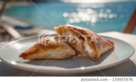 Delicious Greek spinach and feta cheese phyllo pastry triangles, served on a plate with the sparkling blue sea in the background. A close-up of two delicious triangles of Greek spanakopita Delicious Greek spinach and feta cheese phyllo pastry triangles, served on a plate with the sparkling blue sea in the background. A close-up of two delicious triangles of Greek spanakopita 130935676