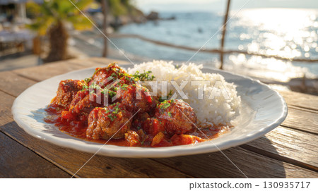 A close-up of a delicious plate of meatballs in tomato sauce with a side of steamed rice, served on a wooden table with the sea in the background. 130935717