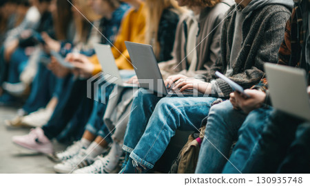 A diverse group of students is seated in a row, each focused on their laptop or mobile device. This image captures essence of education, teamwork, integration of technology in learning environment 130935748