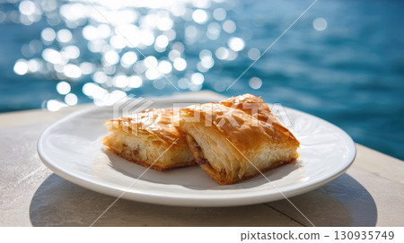 A close-up of two pieces of golden-brown baklava, a traditional Greek pastry, served on a clean white plate. The background features blue ocean with sun glare, creating vacation, summer atmosphere 130935749