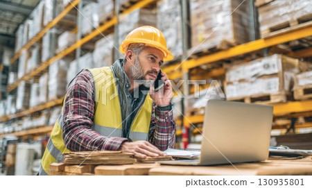 A professional warehouse manager in a hard hat and high-visibility vest is sitting at a desk with a laptop, talking on the phone. This photo represents a busy work day in a logistics hub A professional warehouse manager in a hard hat and high-visibility vest is sitting at a desk with a laptop, talking on the phone. This photo represents a busy work day in a logistics hub 130935801