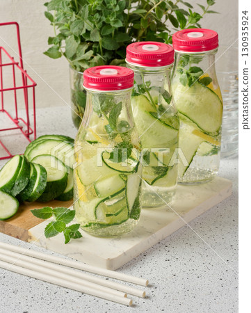 Glass bottles with red lids, filled with detox water containing cucumber and lemon slices and mint leaves 130935924