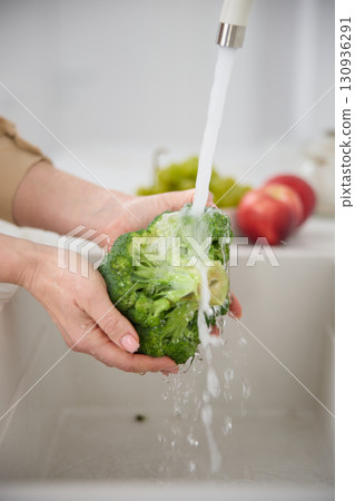 Close up view of a woman washes broccoli under the pressure of flowing water in the white kitchen, tomatoes and green grapes in the background 130936291