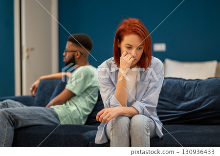 Upset diverse couple wife and husband sit separately on sofa at home not talking after quarrel Upset diverse couple wife and husband sit separately on sofa at home not talking after quarrel 130936433