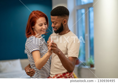 Loving young diverse couple dancing romantic dance in modern bedroom with panoramic windows. 130936495
