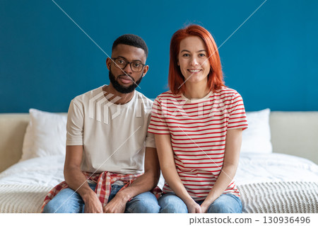 Portrait of happy married interracial diverse couple smiling at camera while sitting together on bed 130936496