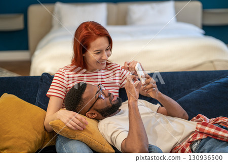 Young diverse couple using smartphone choose hotel for vacation while resting on sofa at home 130936500