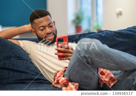 Relaxed smiling african american smiling man sits on couch looks at smartphone at home. Relaxed smiling african american smiling man sits on couch looks at smartphone at home. 130936517