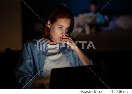 Serious focused woman freelancer working on laptop in dark at home looking at screen.  130936552