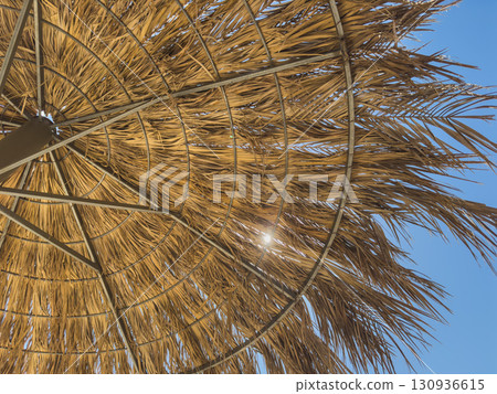 Parasol straw umbrella with sunlight rays. Straw umbrella on hot summer day by beach. Sunny blue skies warm weather enjoyed on beach under parasol made of straw. Vacation in tropical countries 130936615