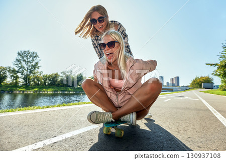 Two women enjoying a skateboard ride on a sunny biking trail 130937108