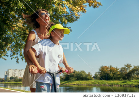 Mother and daughter enjoying a scooter ride along a summer riverside path 130937112