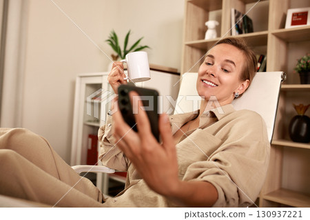 Smiling woman enjoying a coffee while using her smartphone indoors Smiling woman enjoying a coffee while using her smartphone indoors 130937221