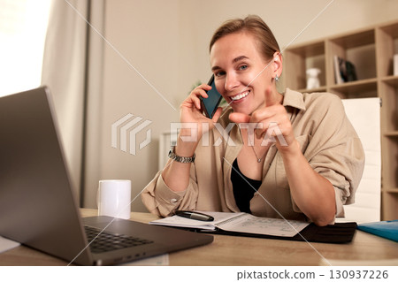 Woman smiling while working on laptop during a phone call at her desk 130937226