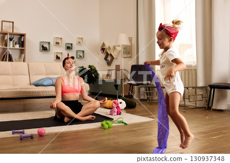 Mother Practicing Yoga While Her Daughter Plays in a Cozy Living Room 130937348