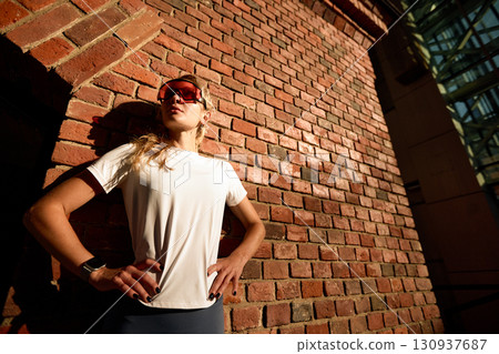 Confident Woman in Athletic Wear Against Red Brick Wall in Urban Setting Confident Woman in Athletic Wear Against Red Brick Wall in Urban Setting 130937687