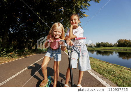 Two cheerful girls playing with their scooters on a sunny outdoor path Two cheerful girls playing with their scooters on a sunny outdoor path 130937735