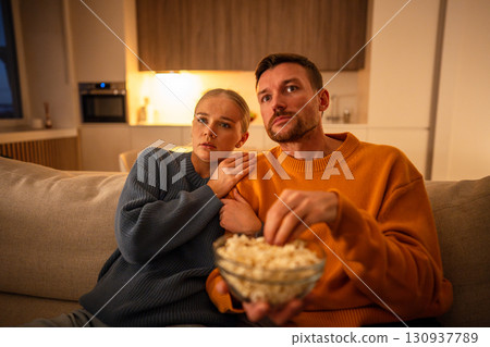 Scared captivated couple eating popcorn watching exciting film on television together in living room Scared captivated couple eating popcorn watching exciting film on television together in living room 130937789