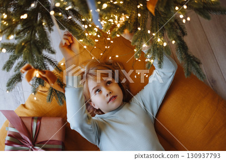 Smiling child top view under Christmas tree with gift box on orange cushion on wooden floor Smiling child top view under Christmas tree with gift box on orange cushion on wooden floor 130937793