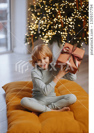 Smiling girl in pajamas tossing gift box near Christmas tree with lights in cozy room Smiling girl in pajamas tossing gift box near Christmas tree with lights in cozy room 130937794