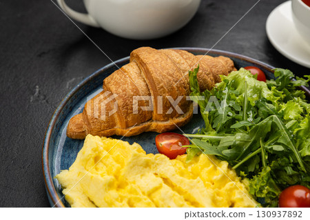 Healthy breakfast with scrambled eggs, croissant, and fresh salad on dark background Healthy breakfast with scrambled eggs, croissant, and fresh salad on dark background 130937892