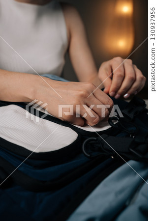 Detail cropped shot of woman packing and unpacking urban backpack sitting in dark living room, organizing essentials for travel, hiking or daily commuting adventures. Concept of casual lifestyle. 130937956