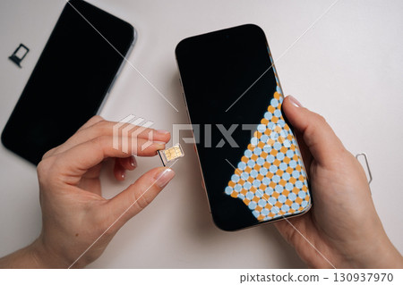 High-angle view of woman inserting sim card into new smartphone, with another phone and sim ejector tool nearby on white table, during mobile device setup process. Concept of modern technology 130937970