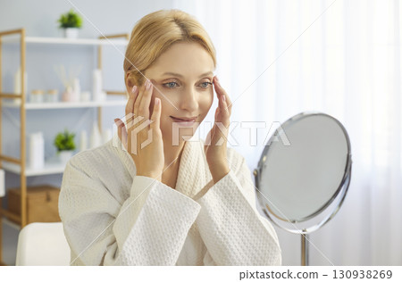 Young blonde woman in bathrobe gently touching her face skin 130938269