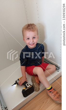 Boy with cordless drill pretends to assemble a cabinet Boy with cordless drill pretends to assemble a cabinet 130938294