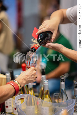 Hand pouring red wine into glass during tasting event, surrounded by bottles and vibrant atmosphere of social gathering. Concept of wine tasting, hospitality, premium experience Hand pouring red wine into glass during tasting event, surrounded by bottles and vibrant atmosphere of social gathering. Concept of wine tasting, hospitality, premium experience 130938492