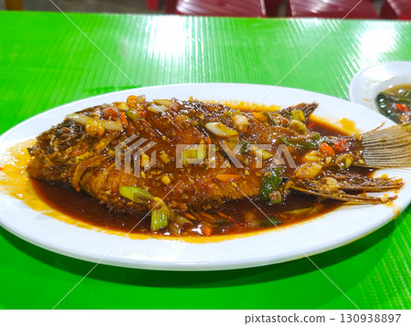 Whole gourami fish served in spicy brown sauce with colorful vegetables on a white plate, set on a green table in a casual Asian restaurant. Whole gourami fish served in spicy brown sauce with colorful vegetables on a white plate, set on a green table in a casual Asian restaurant. 130938897
