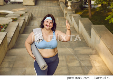 Fat woman in sportswear standing in summer park with yoga mat and towel and showing thumb up sign. Fat woman in sportswear standing in summer park with yoga mat and towel and showing thumb up sign. 130940130