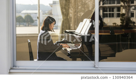 一名女高中生在音樂室彈鋼琴，這是用長焦距鏡頭從校舍外面的窗戶拍攝的 130940364