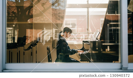 一名女高中生在音樂室彈鋼琴，這是用長焦距鏡頭從校舍外面的窗戶拍攝的 130940372