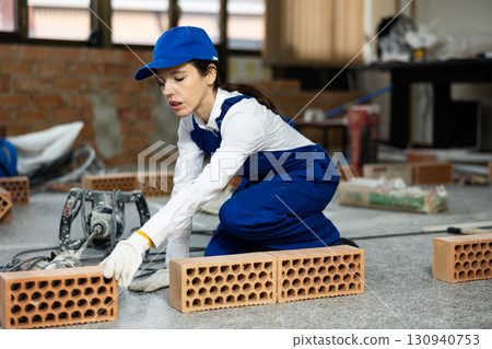 Woman builder in blue overalls lays out bricks on the wall in room 130940753