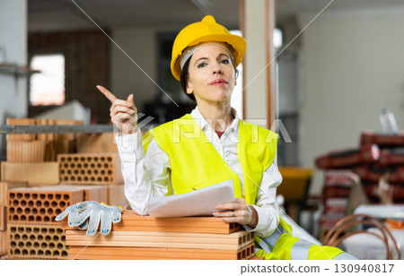 Woman foreman in a yellow vest and a protective helmet points with her hand somewhere in brick warehouse Woman foreman in a yellow vest and a protective helmet points with her hand somewhere in brick warehouse 130940817