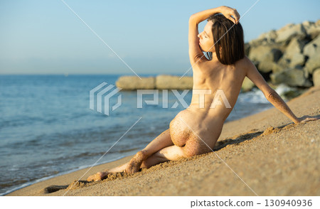 Back view of nude woman sitting on sandy shore in sea waves Back view of nude woman sitting on sandy shore in sea waves 130940936