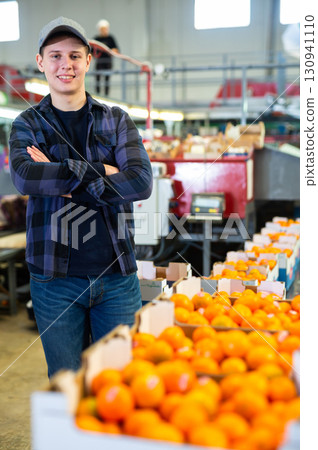 Positive male employee in a warehouse next to boxes of fresh tangerines prepared for sale in a store. Positive male employee in a warehouse next to boxes of fresh tangerines prepared for sale in a store. 130941110