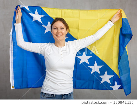 Happy young woman holding flag of Bosnia and Herzegovina against unicoloured background 130941135