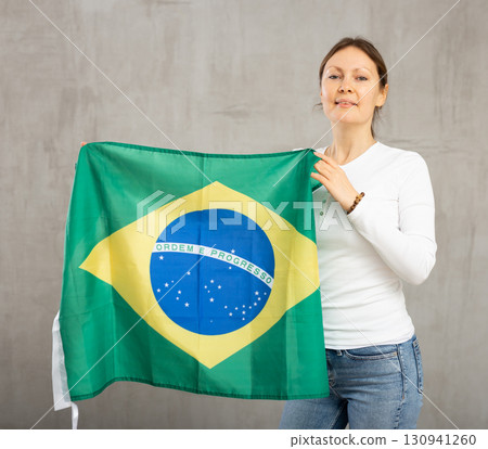 Portrait of confident calm smiling female standing against gray wall with national flag of Brazil 130941260