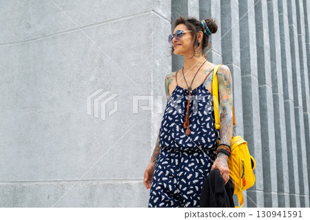 Woman in sunglasses with tattooed arms wearing jumpsuit standing next to a wall. Woman in sunglasses with tattooed arms wearing jumpsuit standing next to a wall. 130941591