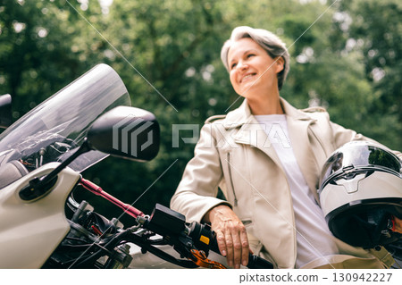 Confident senior woman with gray hair holding helmet by motorcycle, symbol of freedom and vitality 130942227