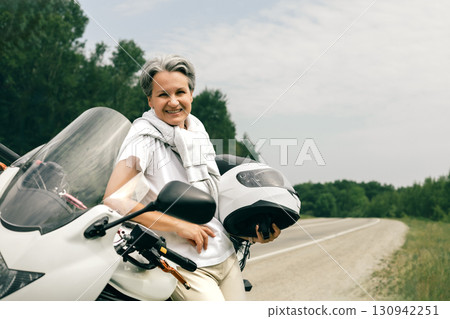 Happy senior woman with gray hair holding helmet near motorbike on rural road, embracing freedom 130942251