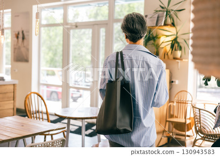 Back view of stylish senior woman entering cozy modern cafe with natural light and minimalist decor 130943583