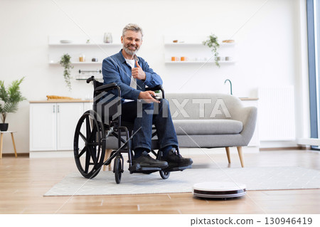 Middle-aged man using robot vacuum cleaner in living room. Clean, organized environment supports assistive lifestyle showcasing independence and technology. Middle-aged man using robot vacuum cleaner in living room. Clean, organized environment supports assistive lifestyle showcasing independence and technology. 130946419