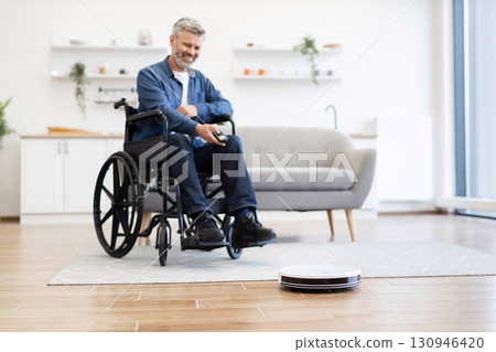 Senior Caucasian man in wheelchair indoors operating robot vacuum cleaner in modern home environment. 130946420