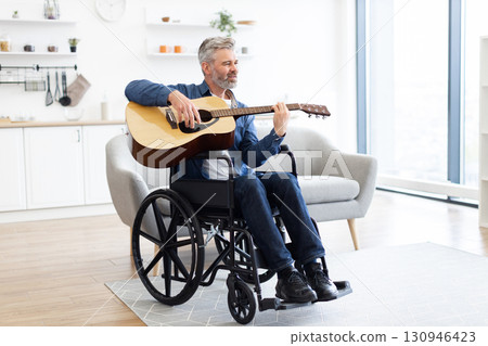 Mature white man playing acoustic guitar sitting in wheelchair indoors in modern living room 130946423