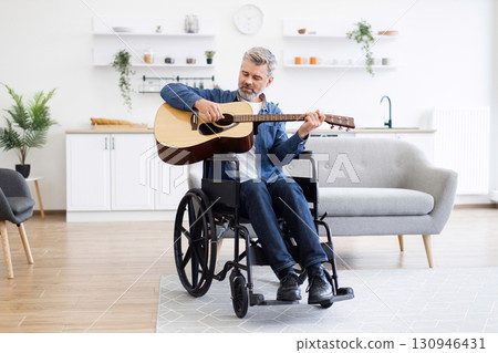 Mature white man playing acoustic guitar in wheelchair in modern living space, expressing music 130946431