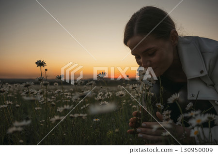 A woman is smelling flowers in a field. The sun is setting, casting a warm glow over the scene. The flowers are scattered throughout the field, with some closer to the woman and others further away. A woman is smelling flowers in a field. The sun is setting, casting a warm glow over the scene. The flowers are scattered throughout the field, with some closer to the woman and others further away. 130950009